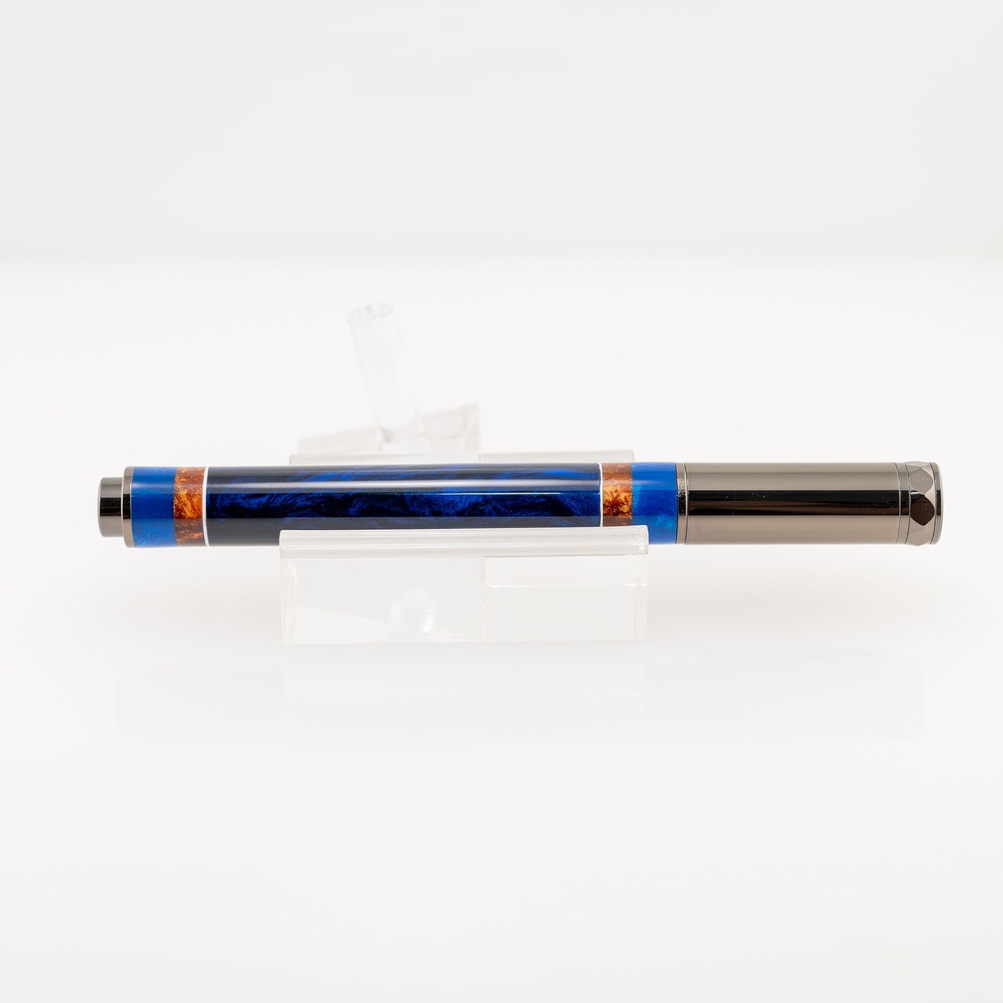 A handmade blue and bronze resin fountain pen rests on a clear stand. The plating is gunmetal and it features a shiny German Iridium nib.