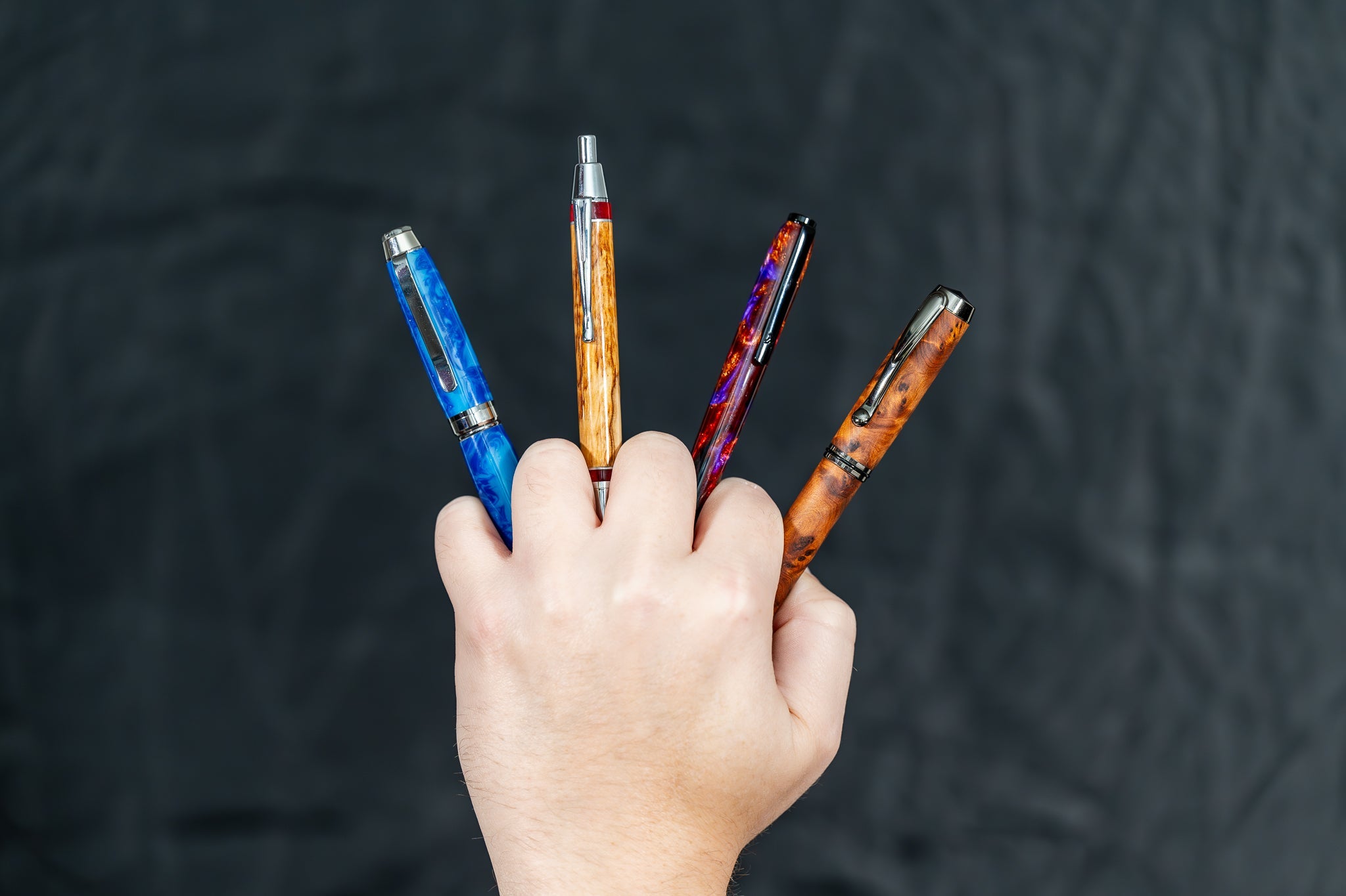 a hand holding four handmade resin, wood, and aluminum pens against a black backgorund
