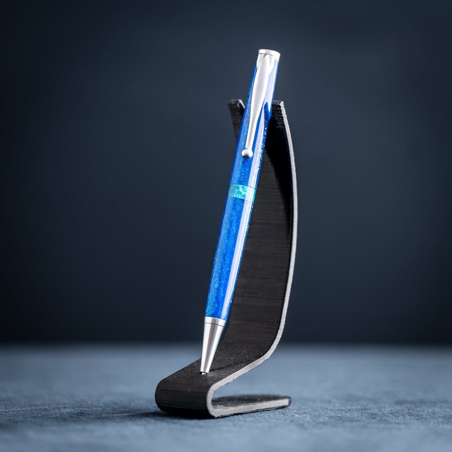 A handmade blue and green resin ballpoint pen with satin chrome plating on a dark stand against a dark background.