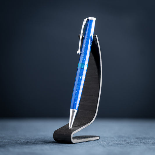 A handmade blue and green resin ballpoint pen with satin chrome plating on a dark stand against a dark background.