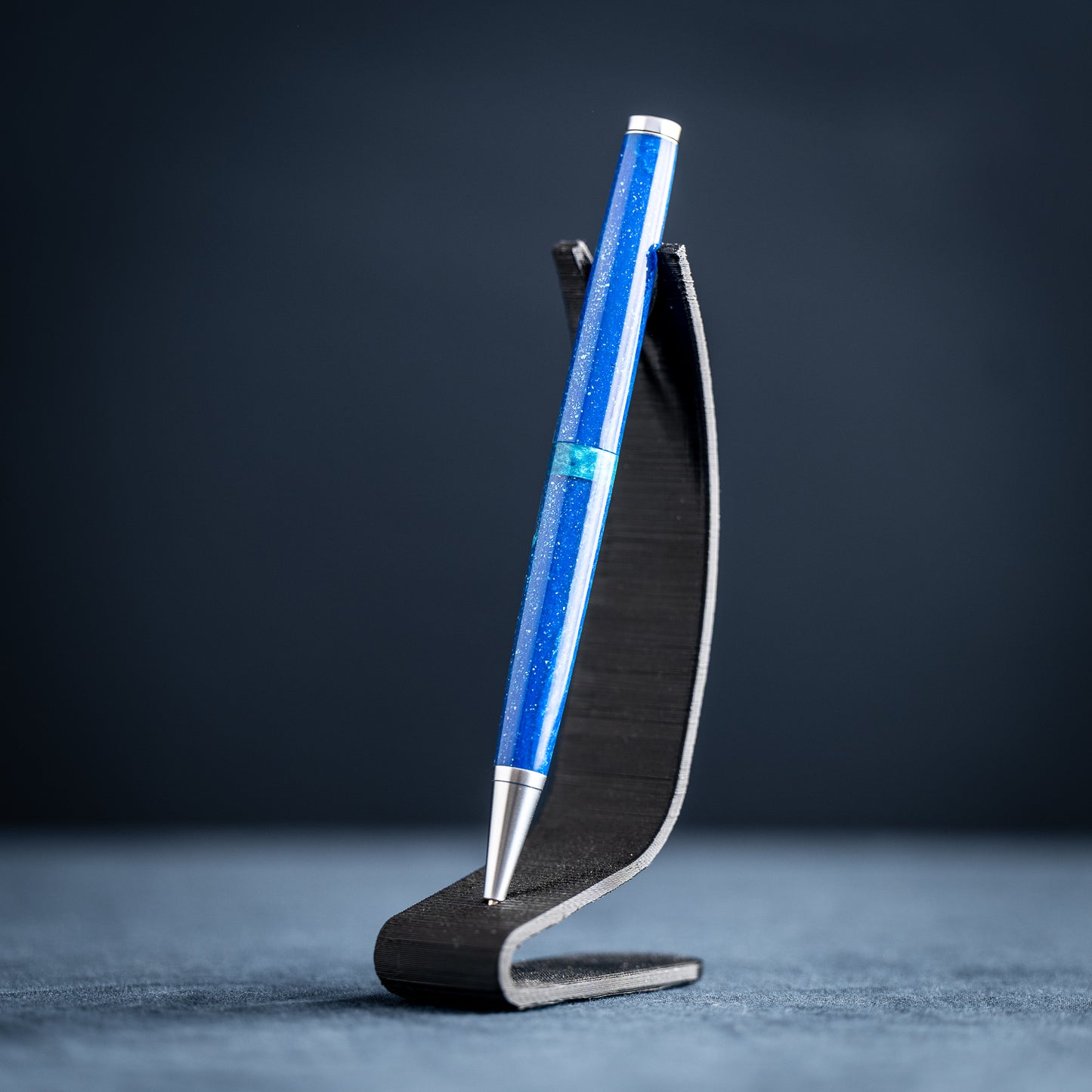 A handmade blue and green resin ballpoint pen with satin chrome plating on a dark stand against a dark background.