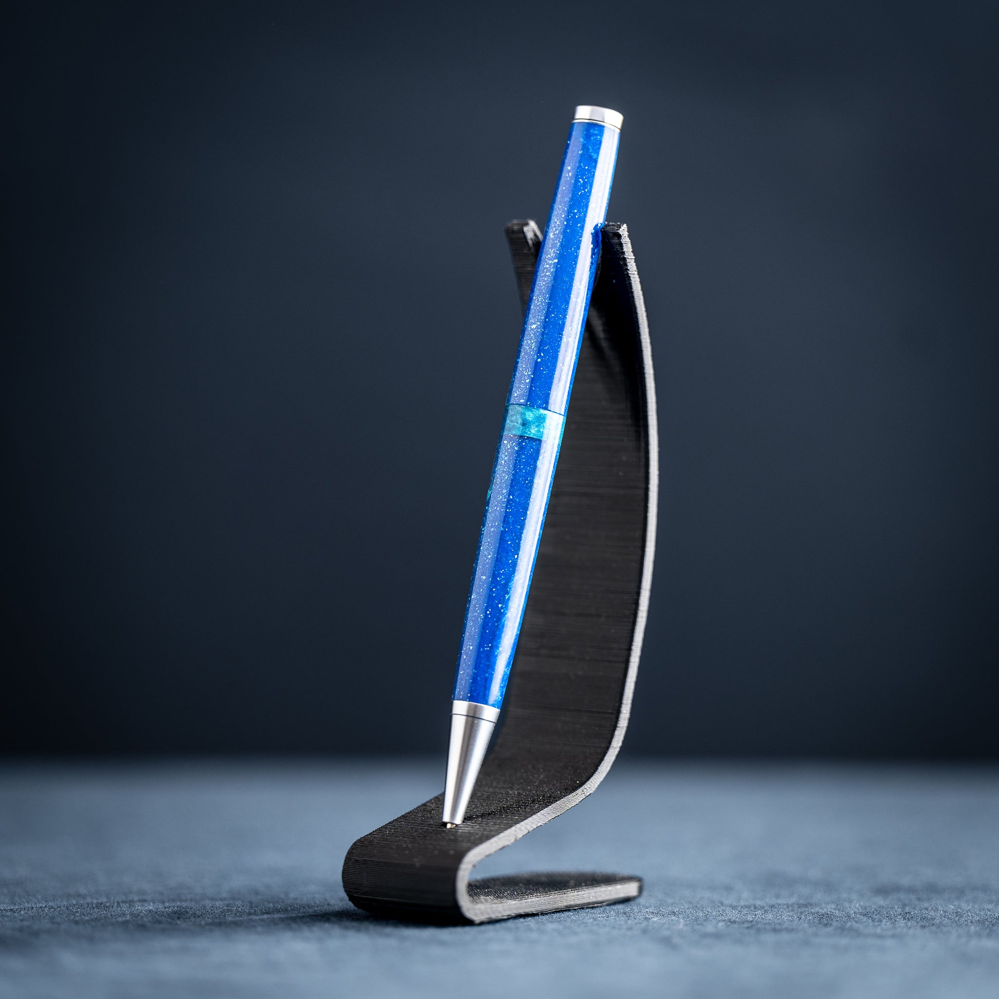 A handmade blue and green resin ballpoint pen with satin chrome plating on a dark stand against a dark background.