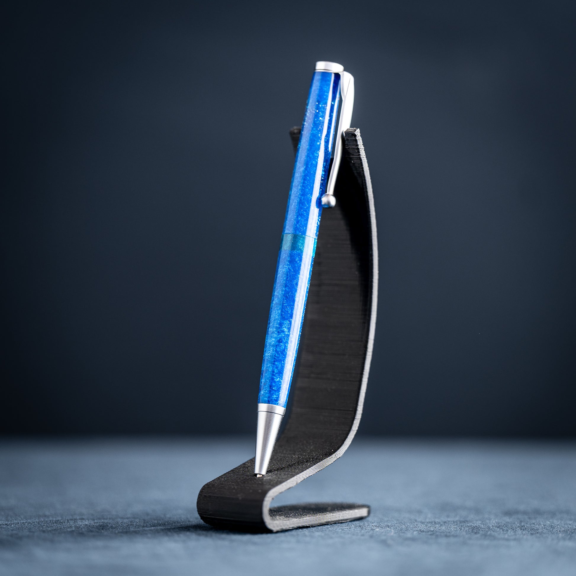 A handmade blue and green resin ballpoint pen with satin chrome plating on a dark stand against a dark background.