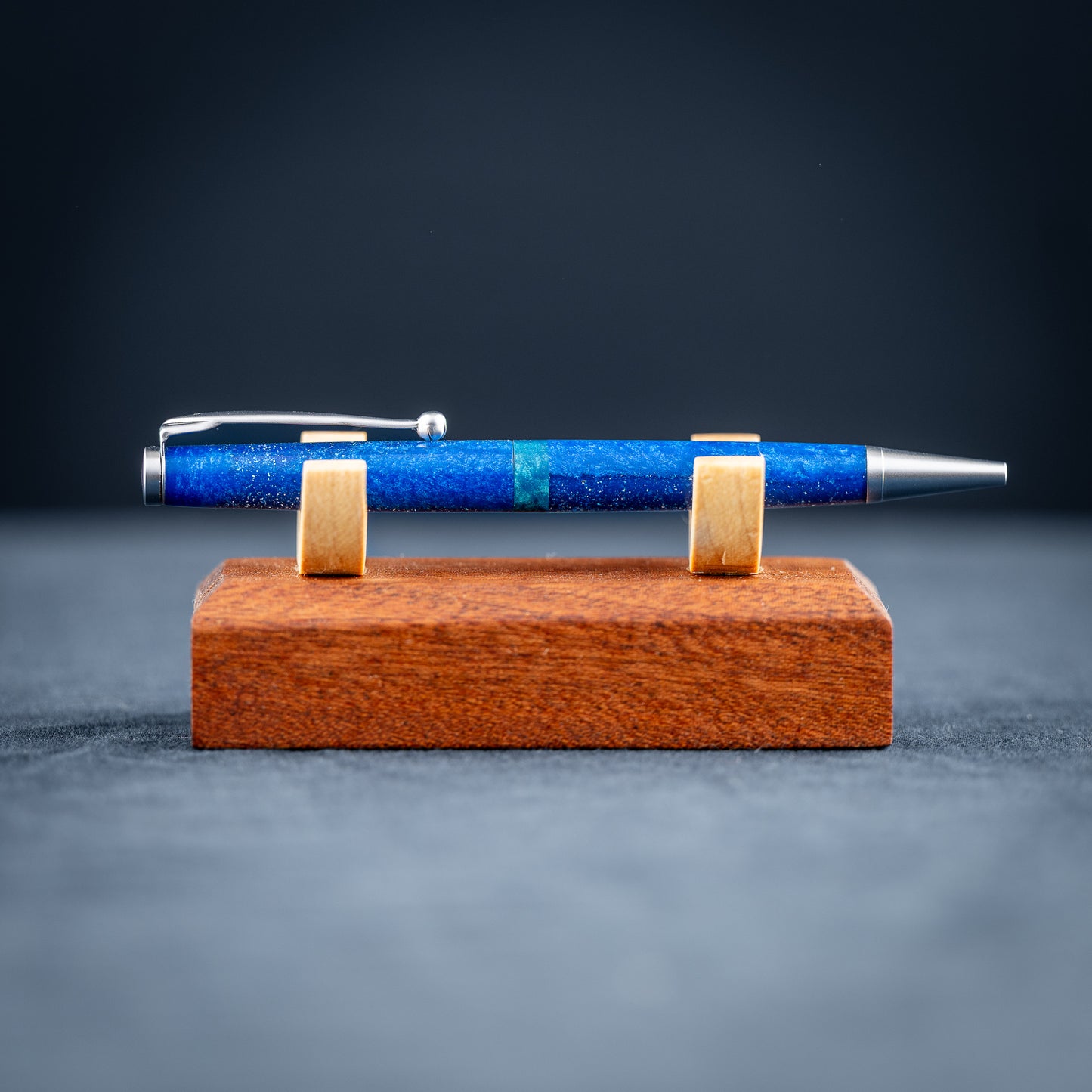 A handmade blue and green resin ballpoint pen with satin chrome plating on a wooden stand against a dark background.