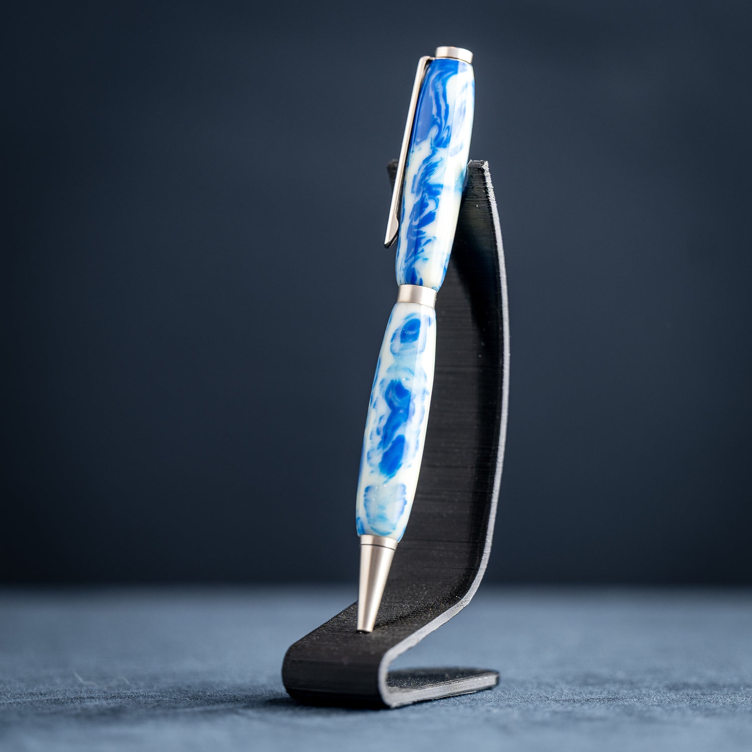 Handmade blue and white resin ballpoint pen with satin chrome plating on a dark stand against a dark background.