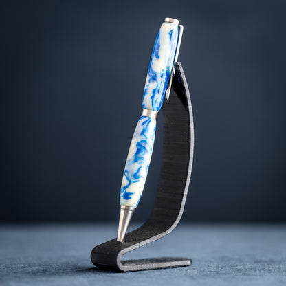 Handmade blue and white resin ballpoint pen with satin chrome plating on a dark stand against a dark background.