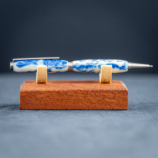 Handmade blue and white resin ballpoint pen with satin chrome plating on a wood stand against a dark background.