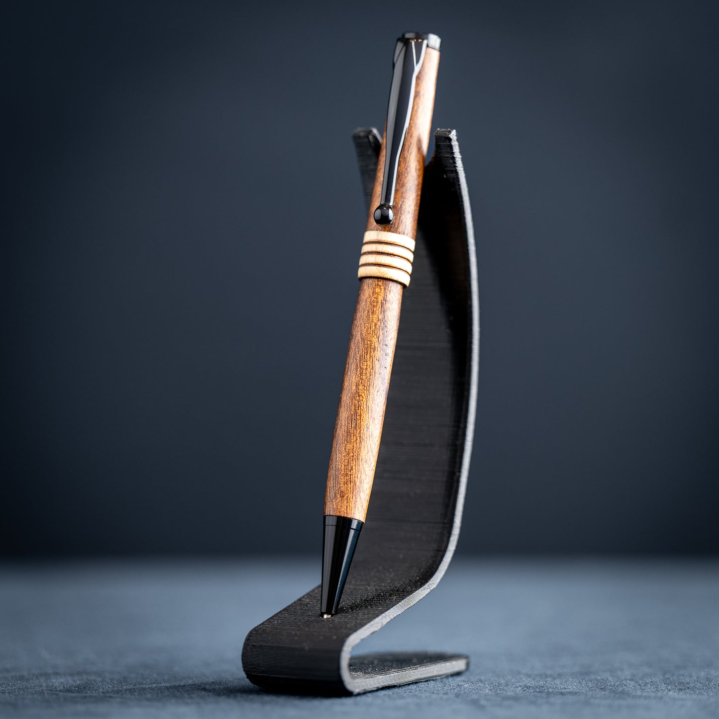 Goncalo alves and maple wood ballpoint pen with black plating on a dark stand against a dark background.