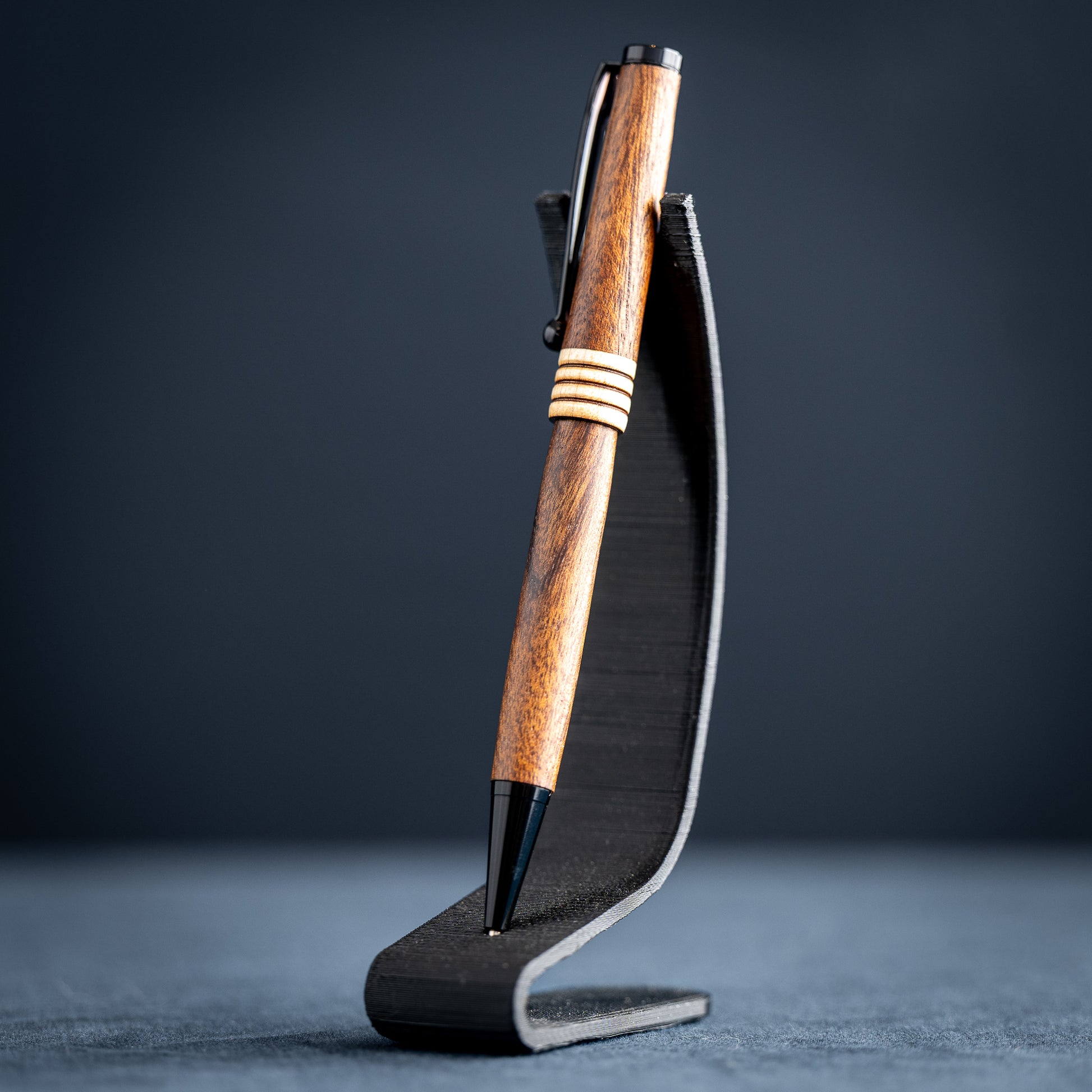Goncalo alves and maple wood ballpoint pen with black plating on a dark stand against a dark background.