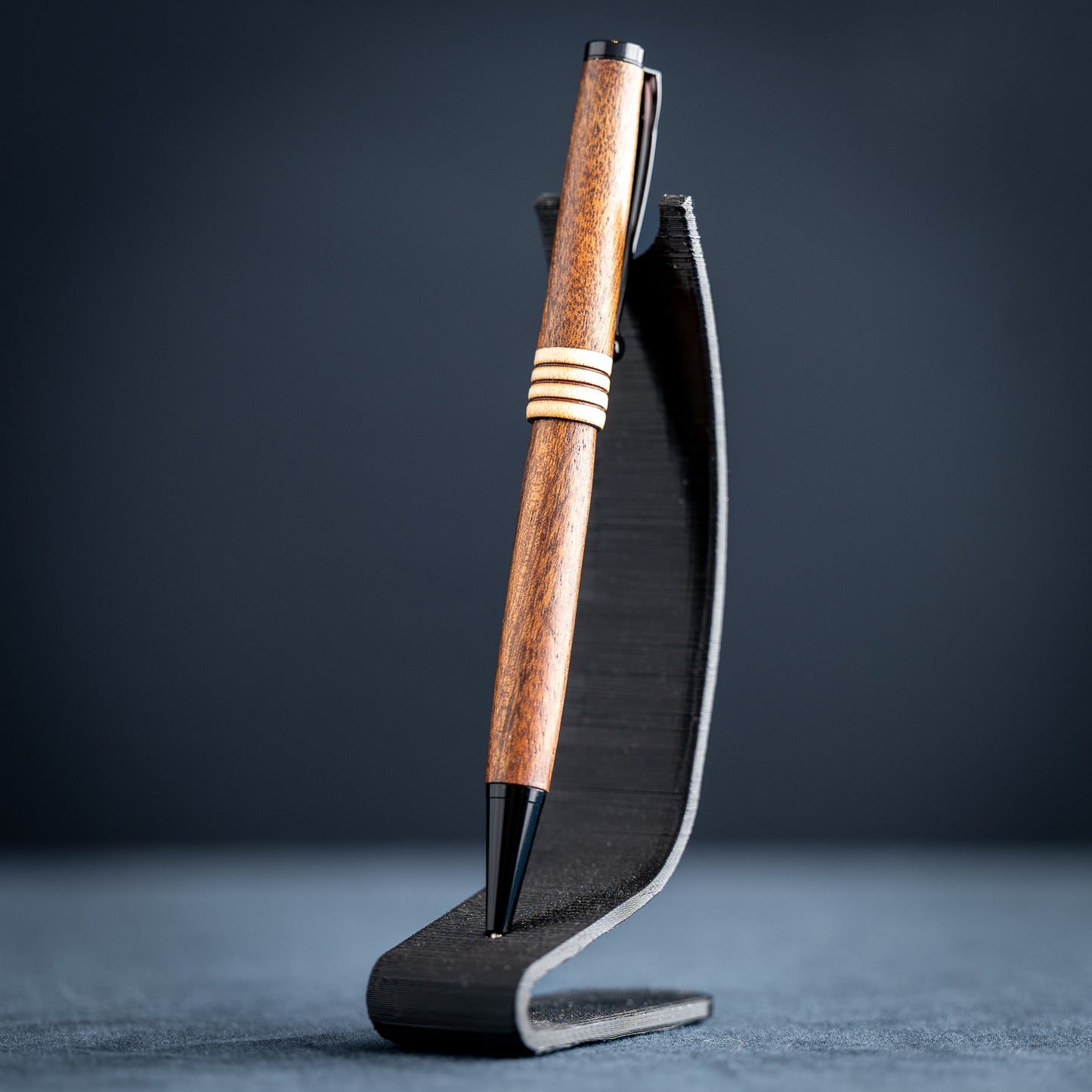 Goncalo alves and maple wood ballpoint pen with black plating on a dark stand against a dark background.
