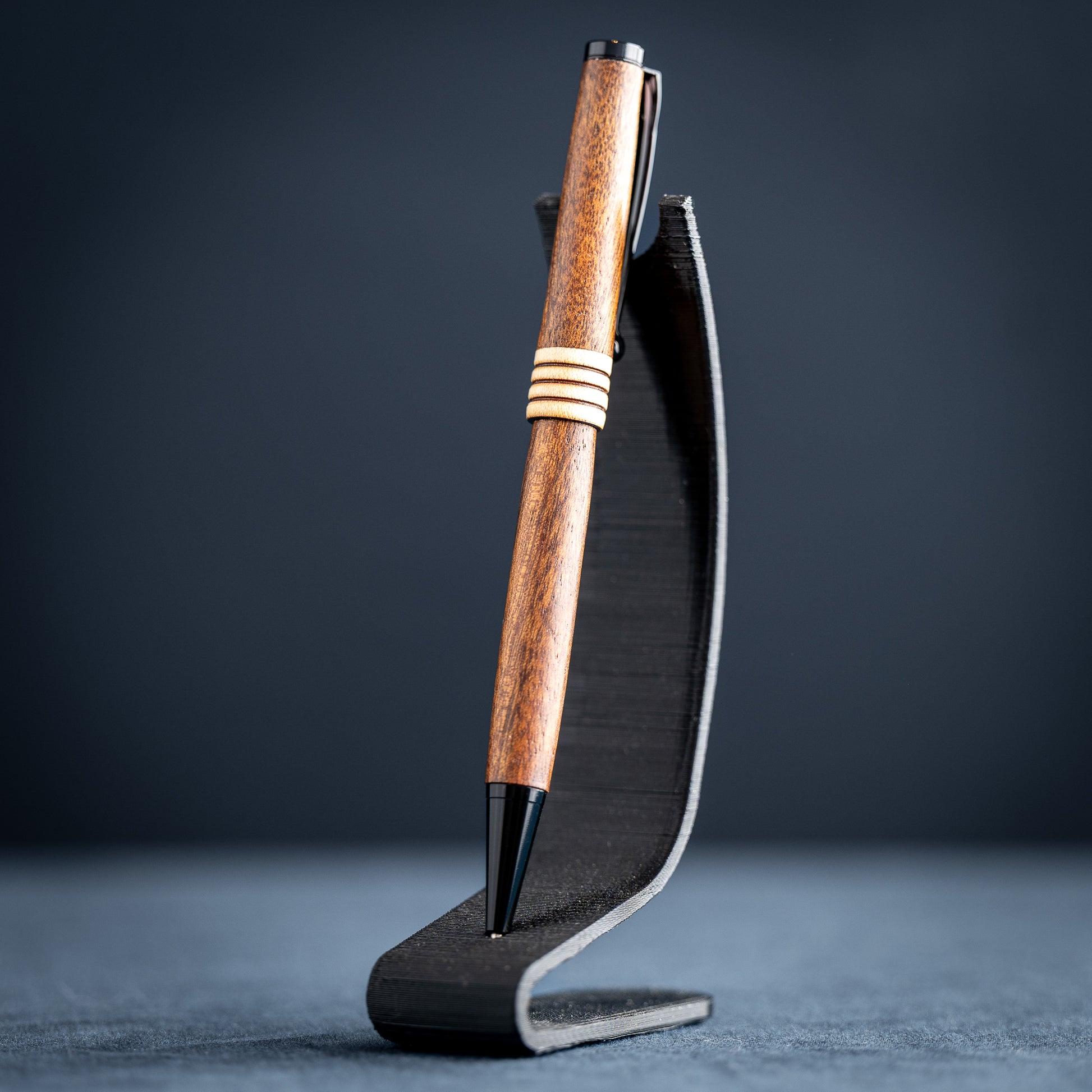 Goncalo alves and maple wood ballpoint pen with black plating on a dark stand against a dark background.
