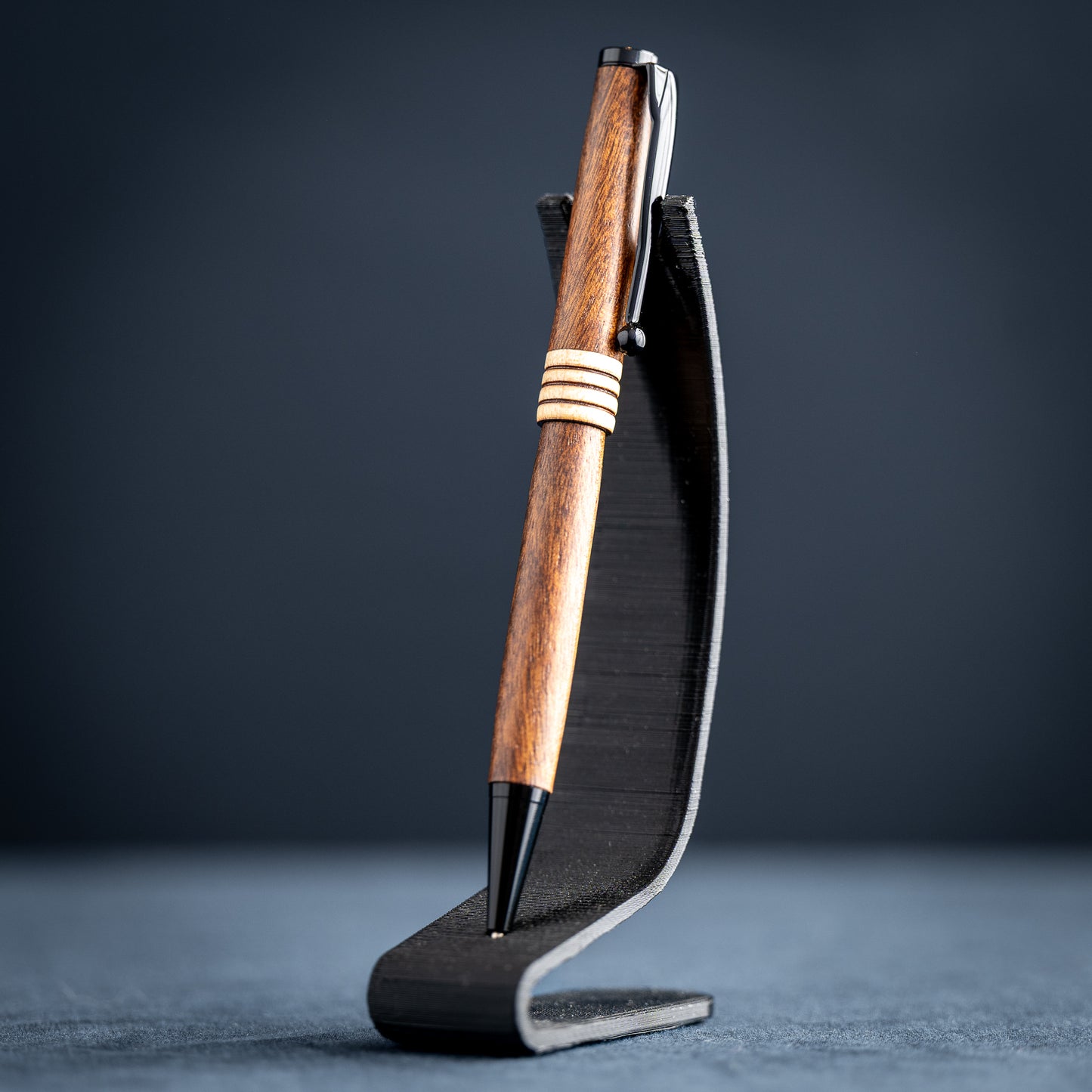 Goncalo alves and maple wood ballpoint pen with black plating on a dark stand against a dark background.