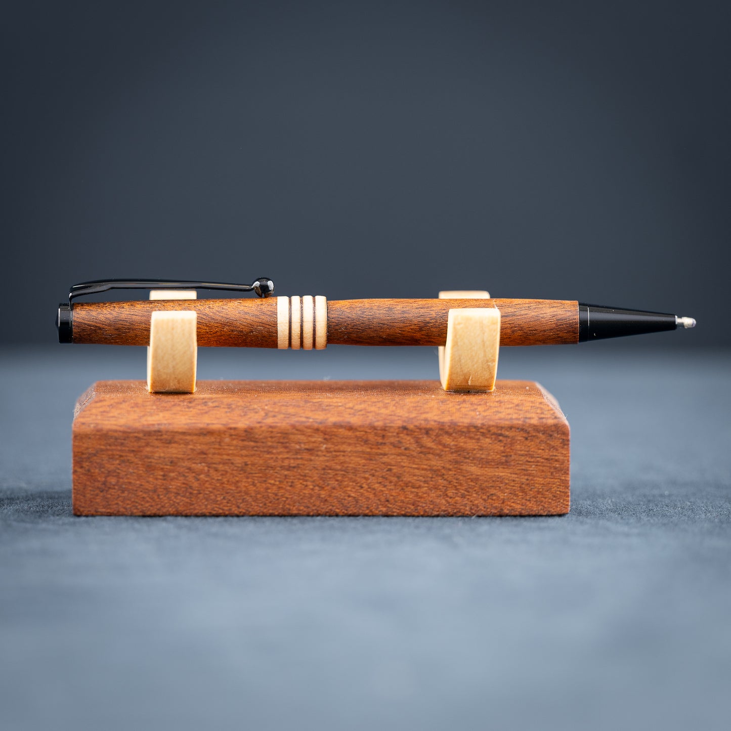 Goncalo alves and maple wood ballpoint pen with black plating on a dark stand against a dark background.