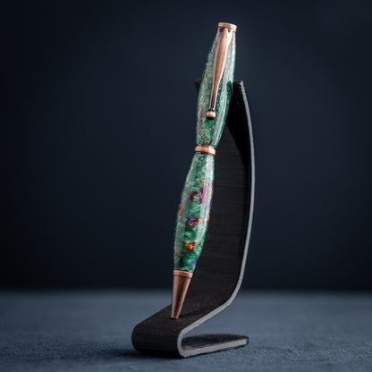 A handmade green, purple, and bronze resin ballpoint twist pen with antique copper plating on a dark stand against a dark background.