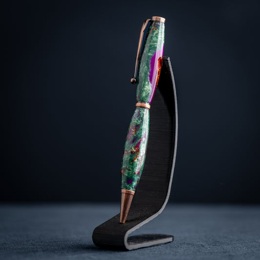 A handmade green, purple, and bronze resin ballpoint twist pen with antique copper plating on a dark stand against a dark background.