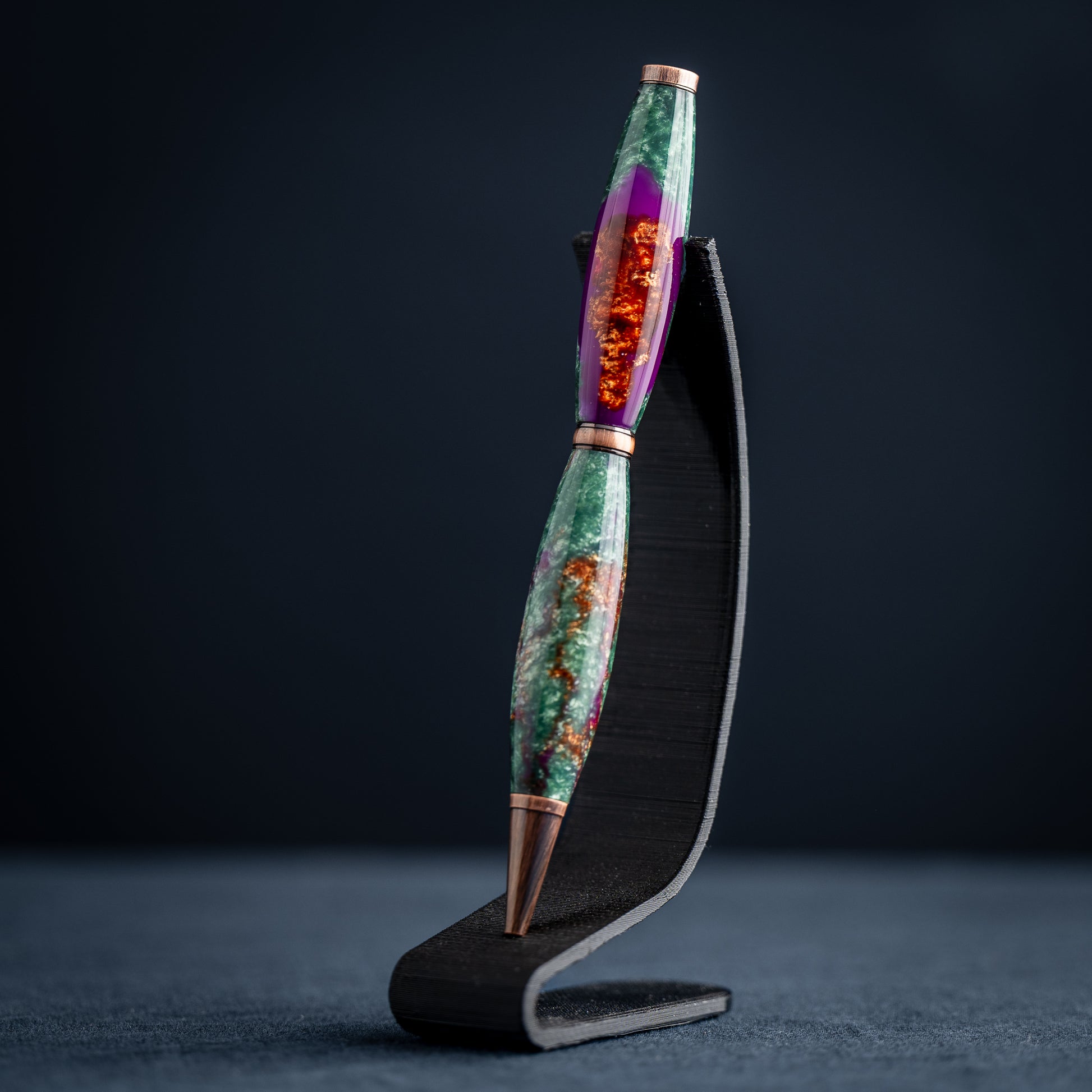 A handmade green, purple, and bronze resin ballpoint twist pen with antique copper plating on a dark stand against a dark background.