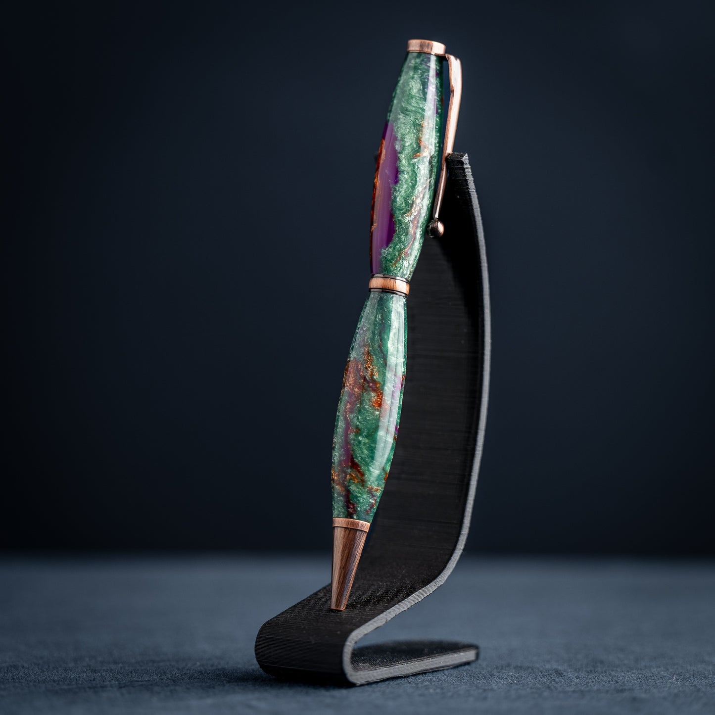 A handmade green, purple, and bronze resin ballpoint twist pen with antique copper plating on a dark stand against a dark background.