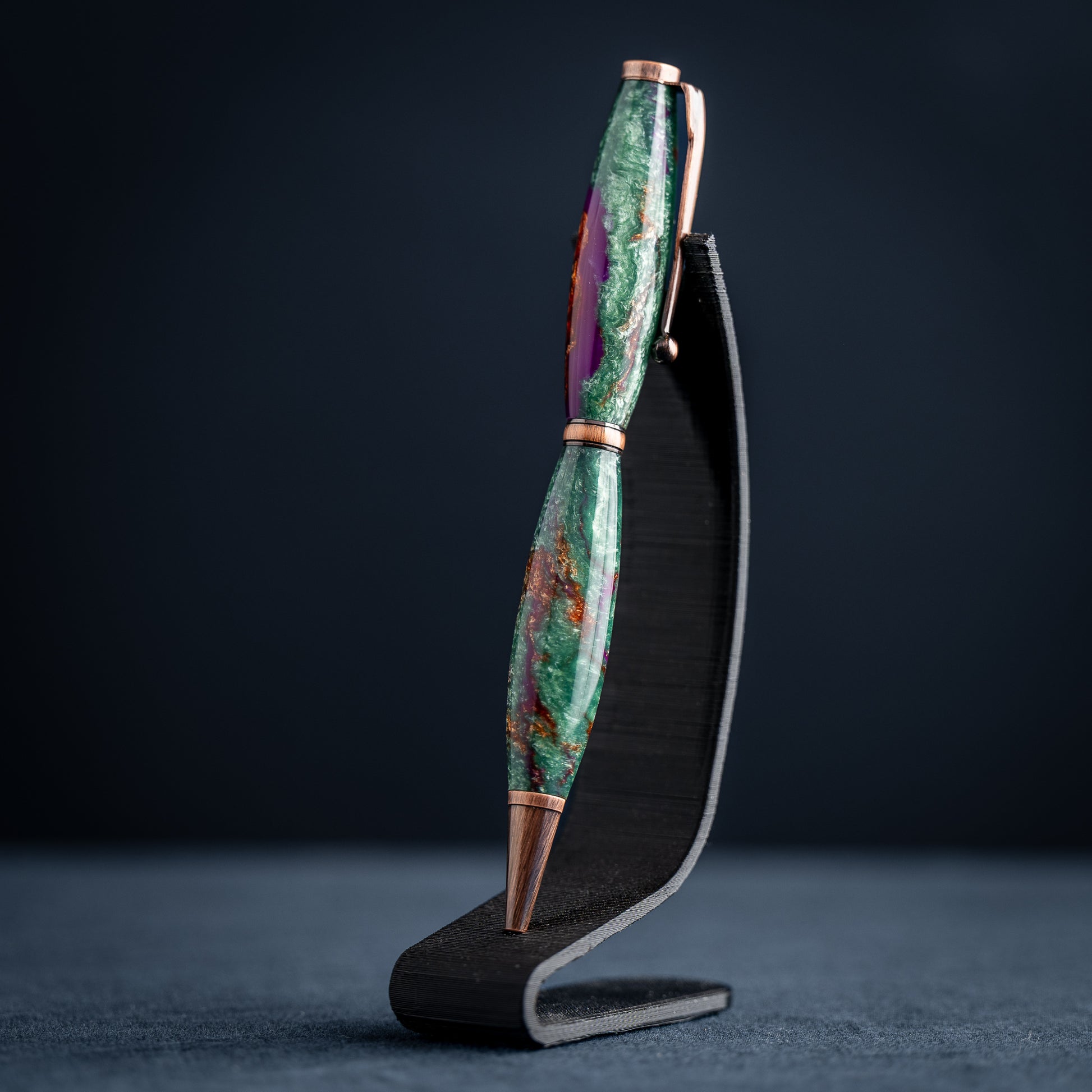 A handmade green, purple, and bronze resin ballpoint twist pen with antique copper plating on a dark stand against a dark background.