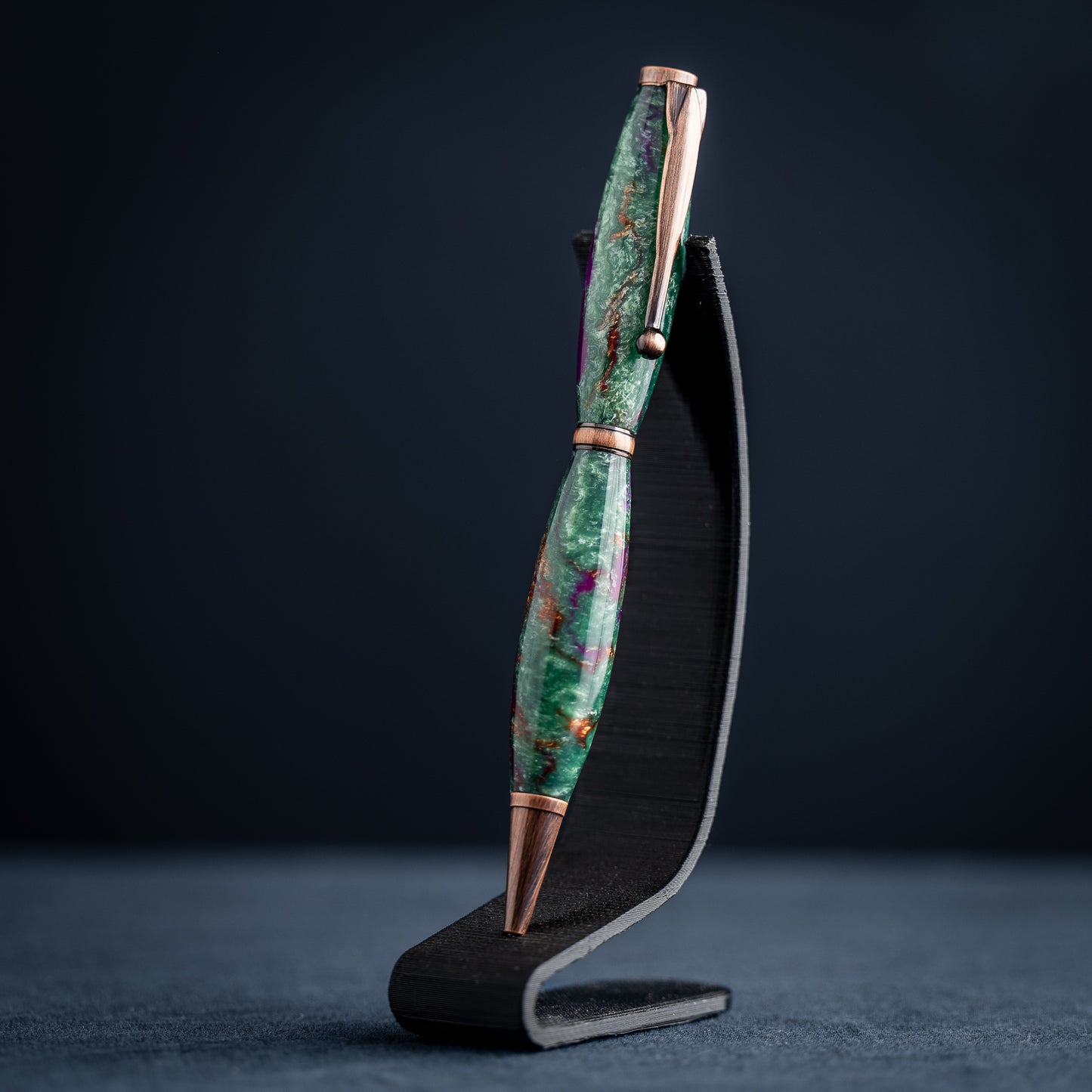 A handmade green, purple, and bronze resin ballpoint twist pen with antique copper plating on a dark stand against a dark background.
