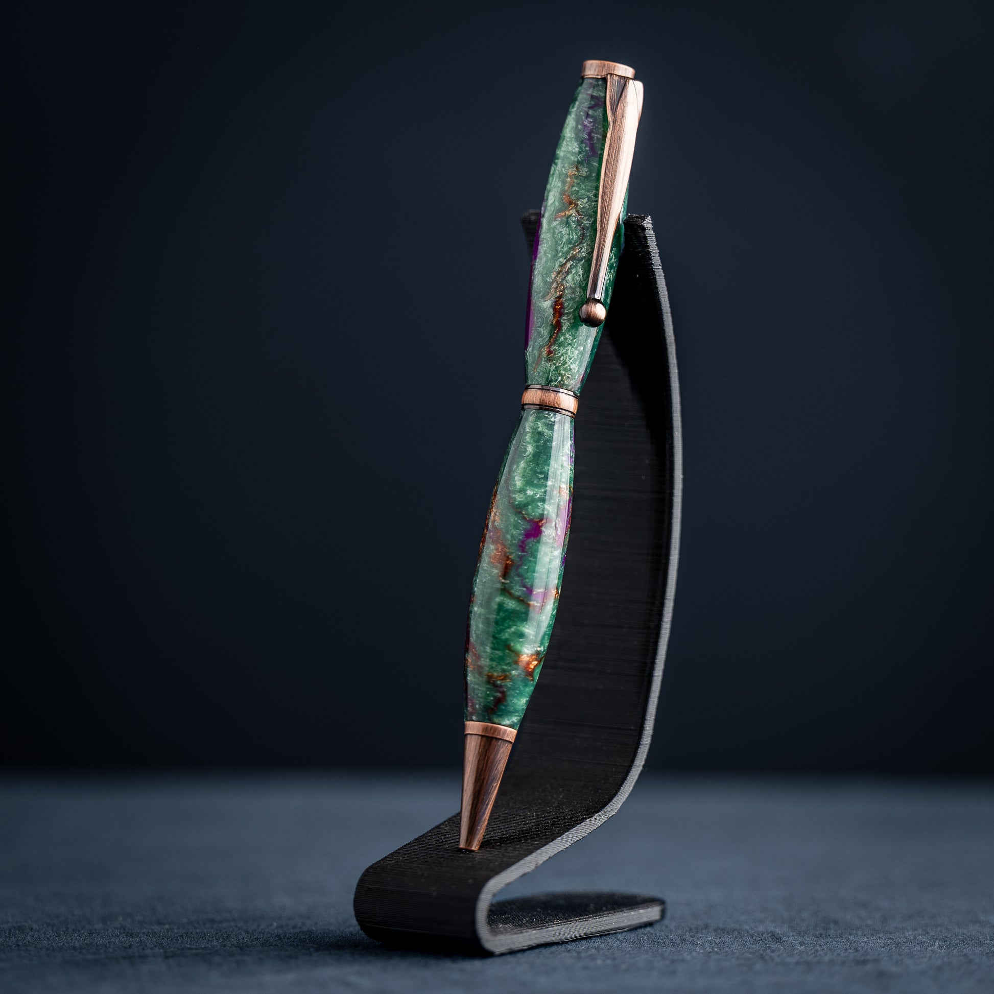 A handmade green, purple, and bronze resin ballpoint twist pen with antique copper plating on a dark stand against a dark background.