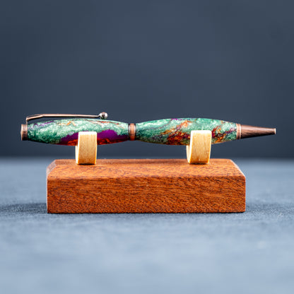 A handmade green, purple, and bronze resin ballpoint twist pen with antique copper plating on a wooden stand against a dark background.