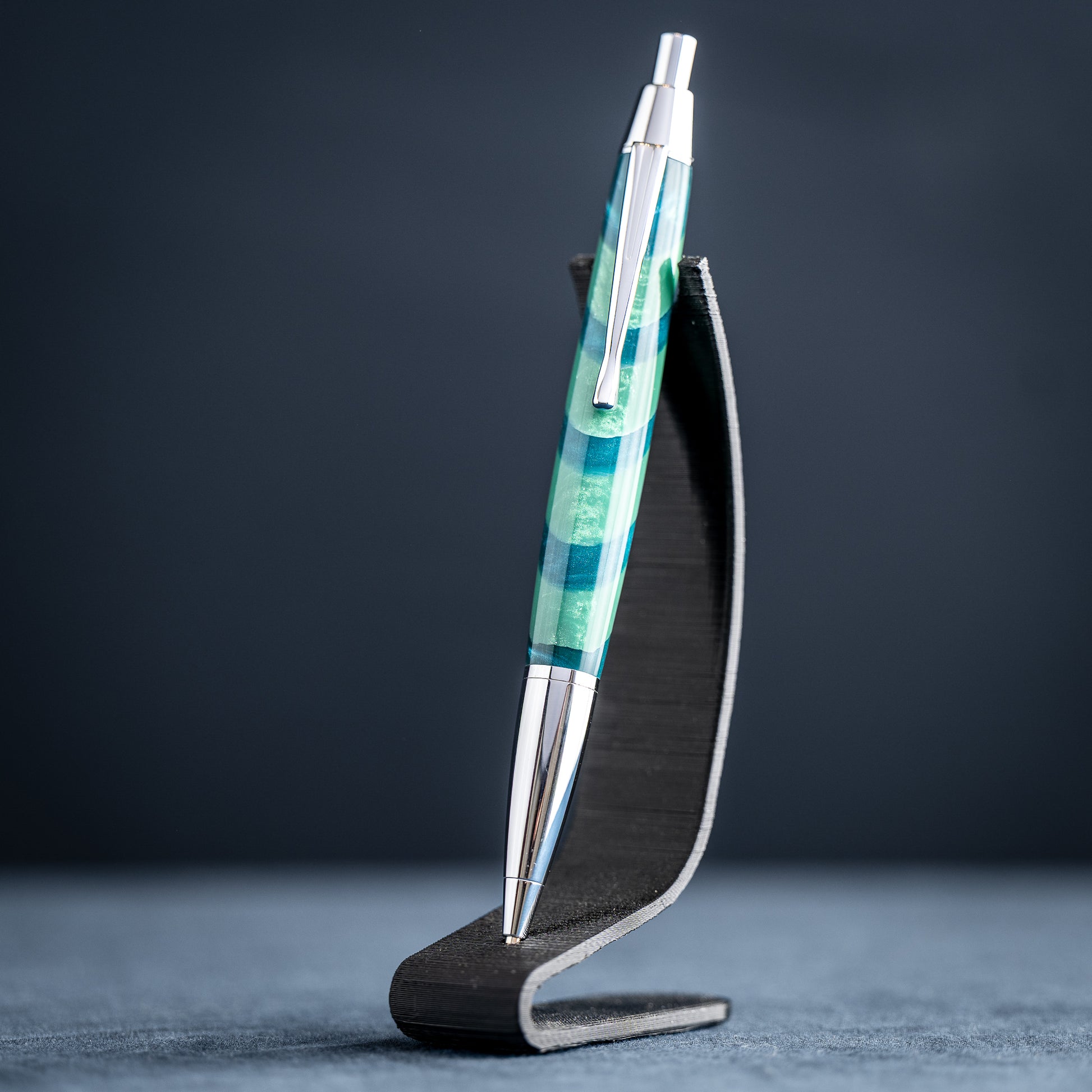 Handmade green striped resin pen with chrome plating on a dark stand against a dark background.