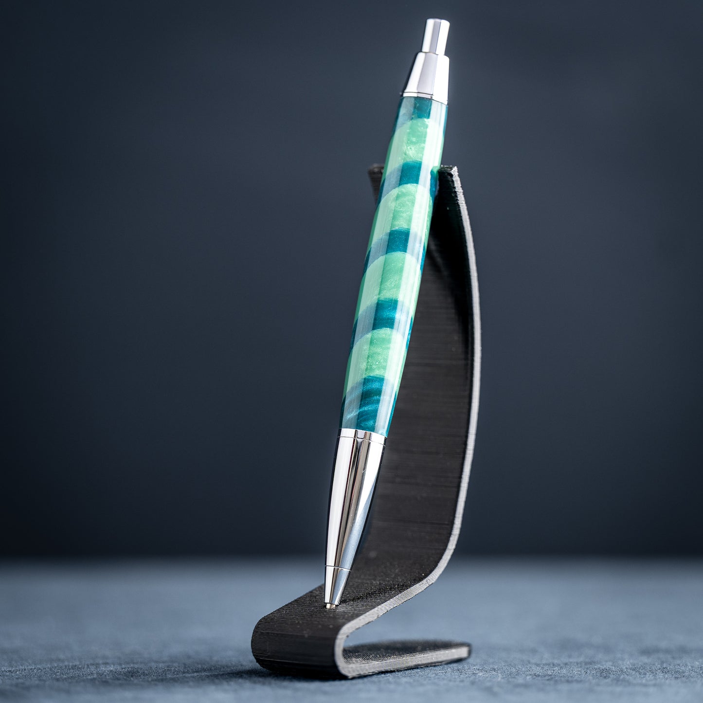 Handmade green striped resin pen with chrome plating on a dark stand against a dark background.