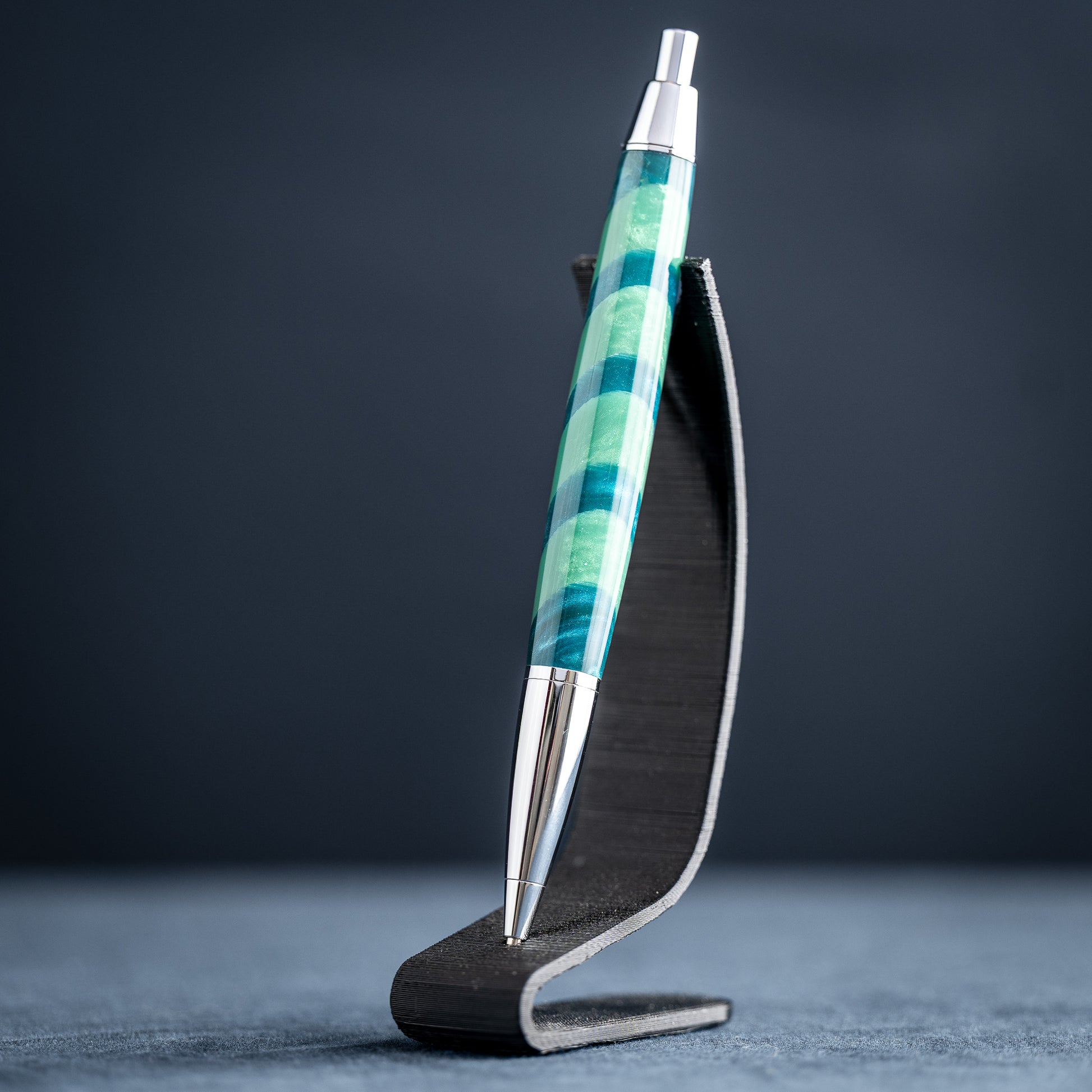 Handmade green striped resin pen with chrome plating on a dark stand against a dark background.