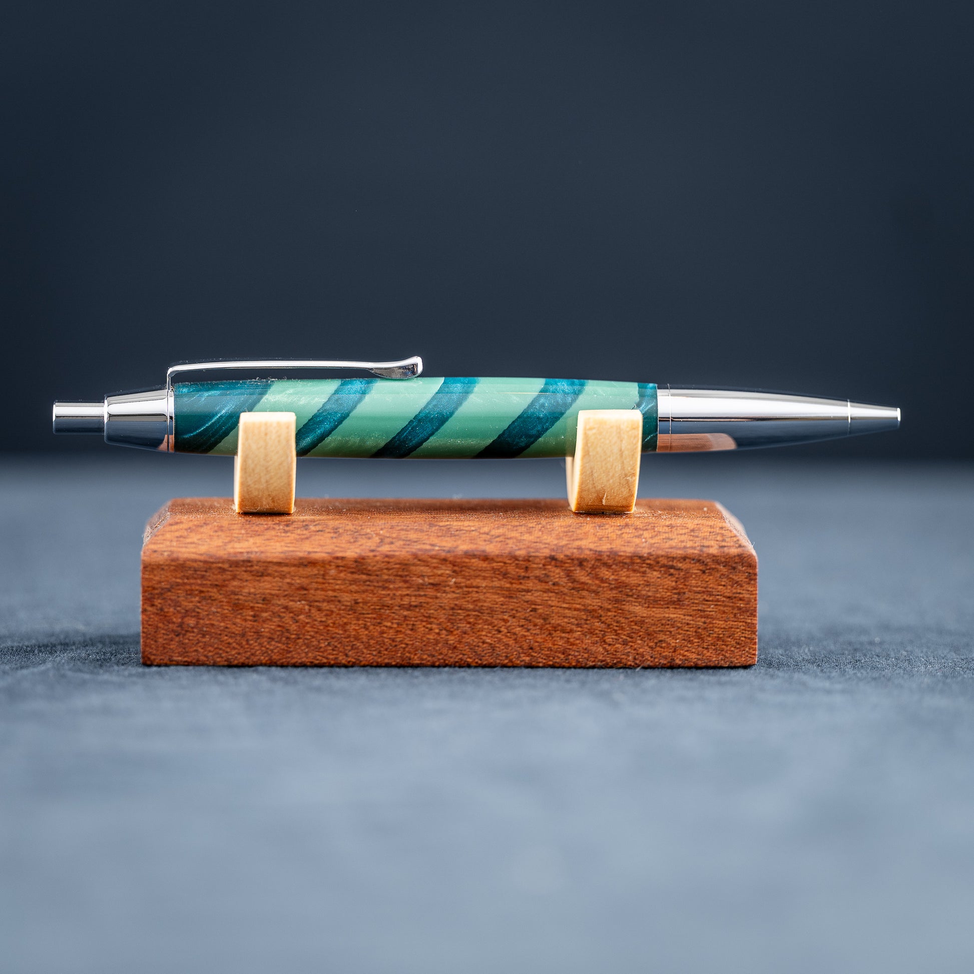 Handmade green striped resin pen with chrome plating on a wooden stand against a dark background.