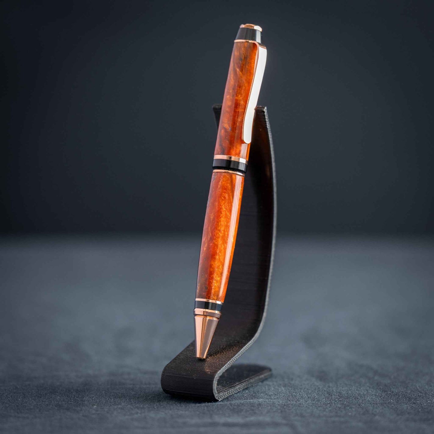 A handmade metallic orange and blue resin twist ballpoint pen with bright copper plating on a dark stand against a dark background.