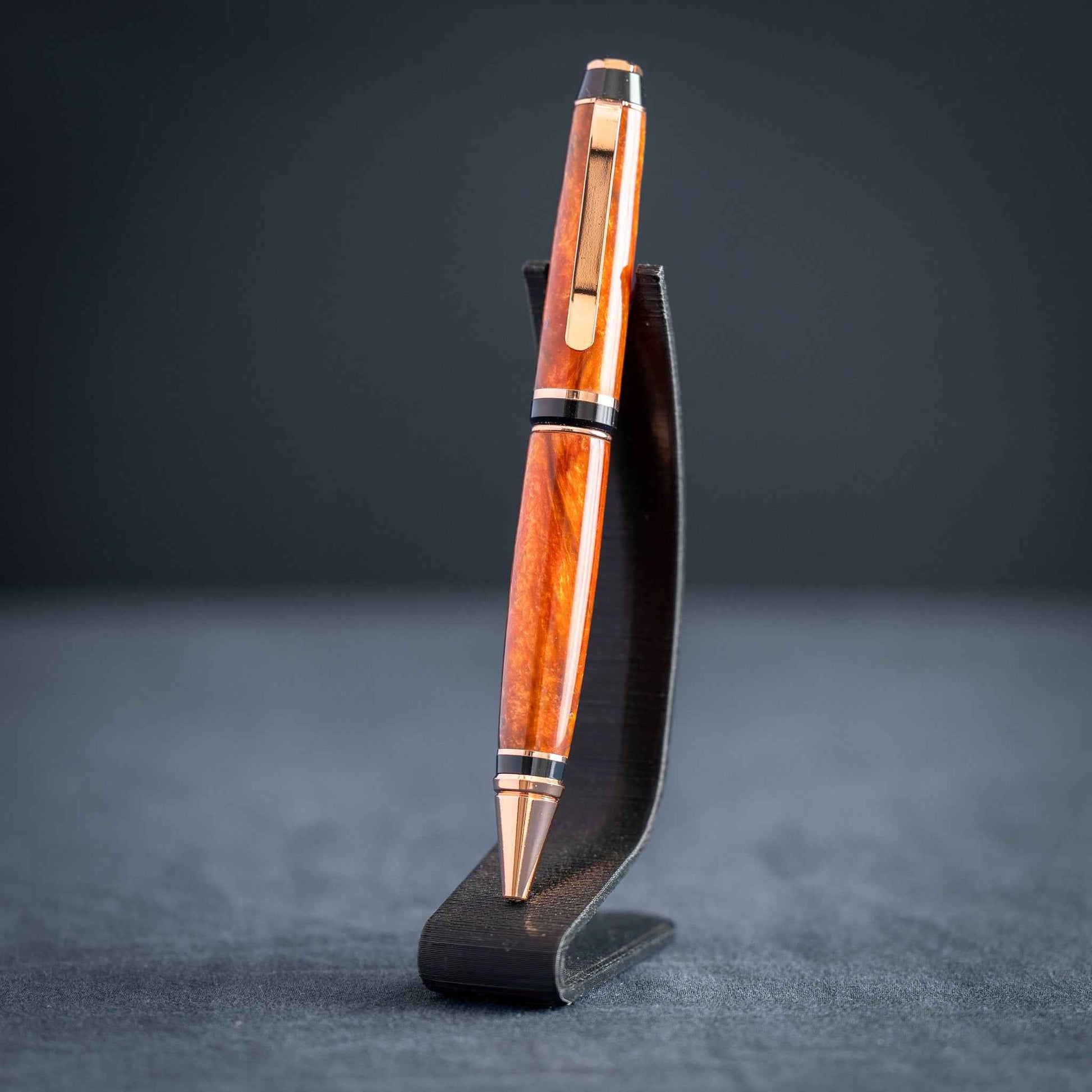 A handmade metallic orange and blue resin twist ballpoint pen with bright copper plating on a dark stand against a dark background.