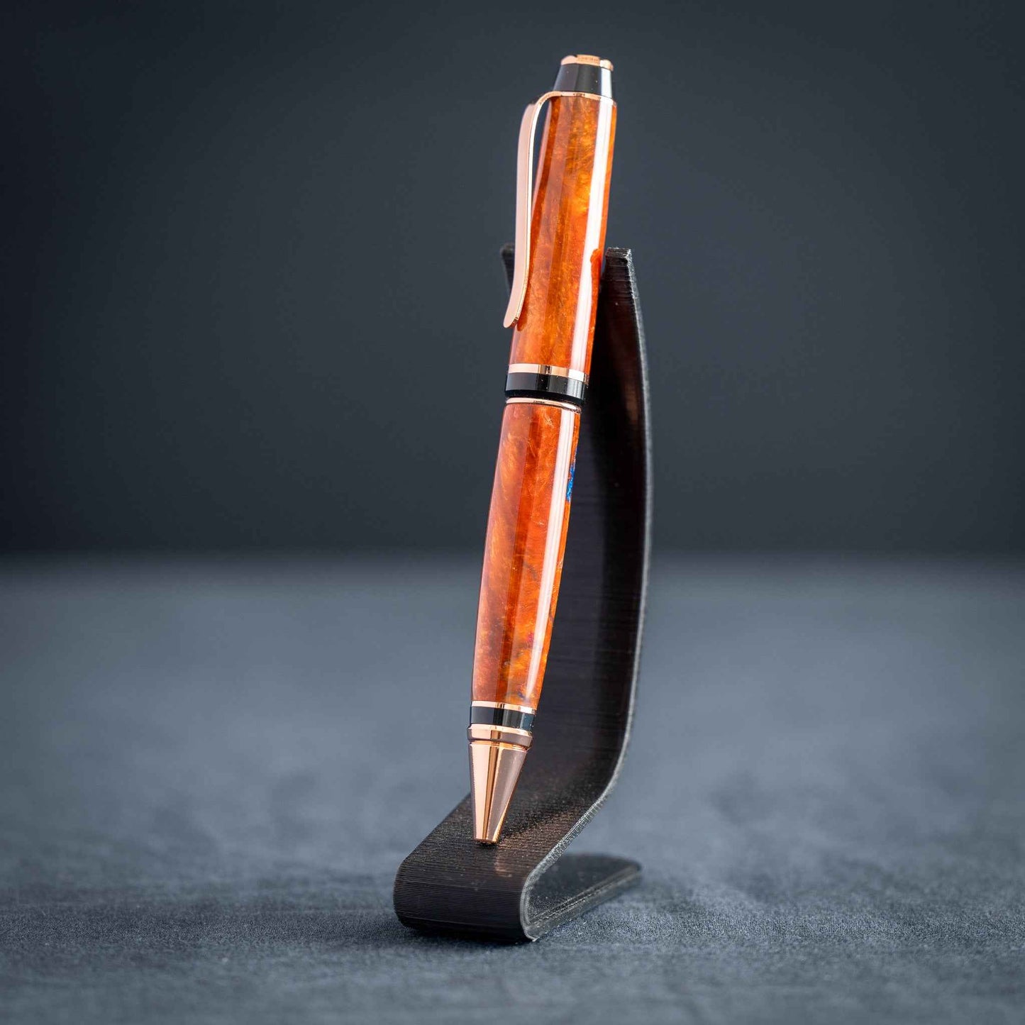 A handmade metallic orange and blue resin twist ballpoint pen with bright copper plating on a dark stand against a dark background.