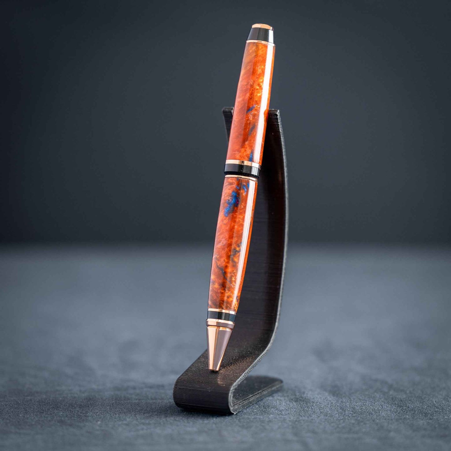 A handmade metallic orange and blue resin twist ballpoint pen with bright copper plating on a dark stand against a dark background.