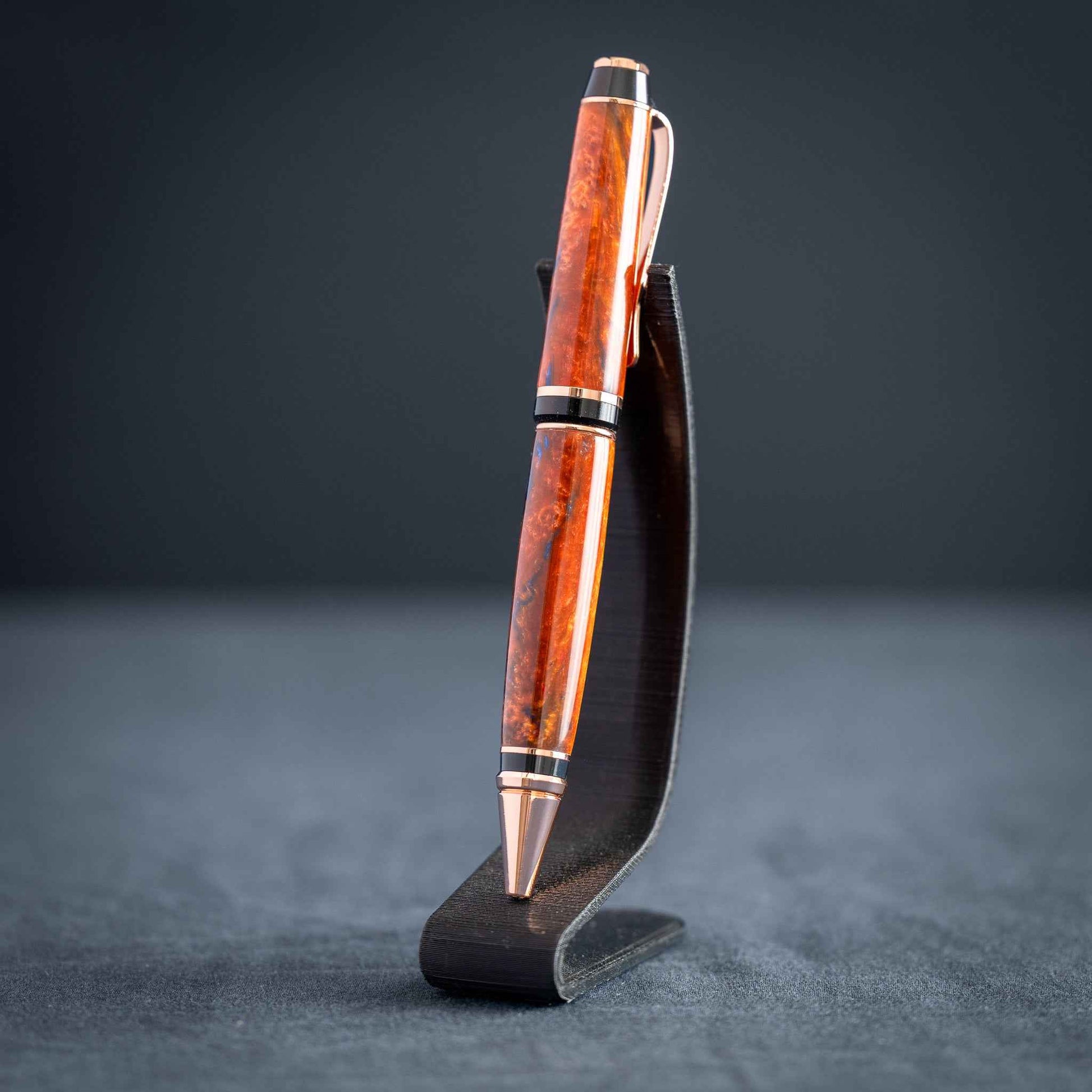 A handmade metallic orange and blue resin twist ballpoint pen with bright copper plating on a dark stand against a dark background.