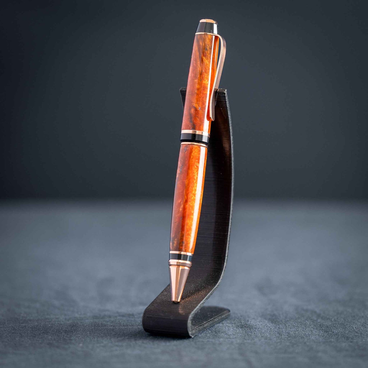 A handmade metallic orange and blue resin twist ballpoint pen with bright copper plating on a dark stand against a dark background.