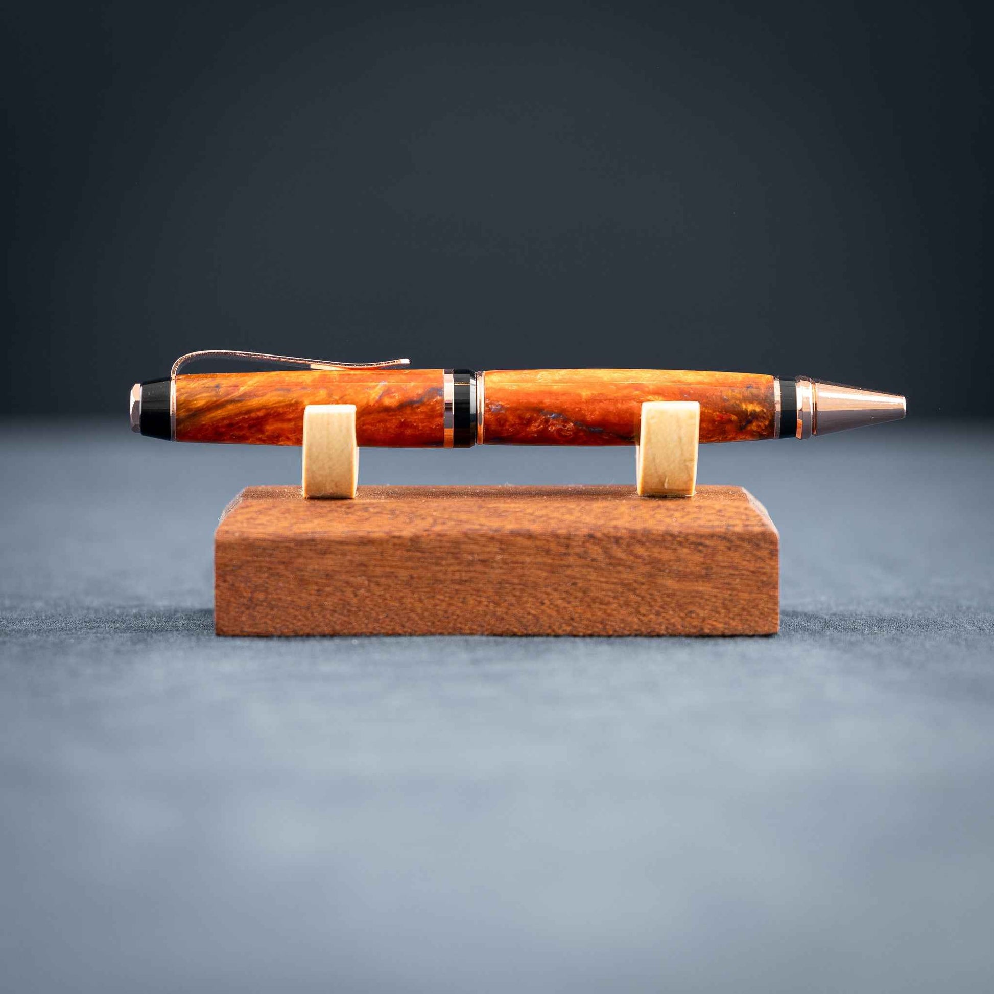 A handmade metallic orange and blue resin twist ballpoint pen with bright copper plating on a dark stand against a dark background.