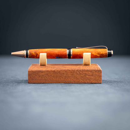 A handmade metallic orange and blue resin twist ballpoint pen with bright copper plating on a dark stand against a dark background.