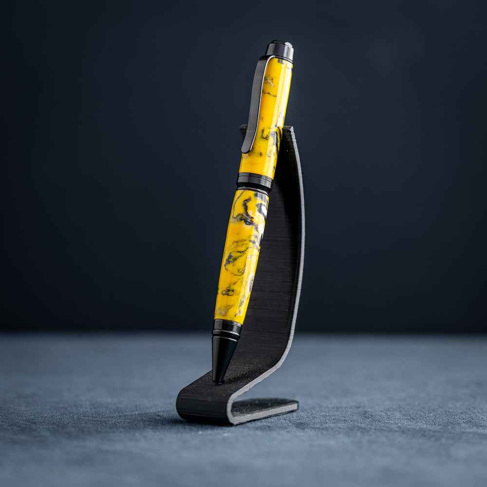 A handmade black, yellow, and white resin ballpoint pen on a black stand against a black background.