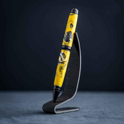 A handmade black, yellow, and white resin ballpoint pen on a wooden stand against a black background.