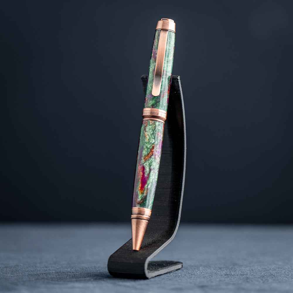 A handmade green, purple, and copper resin ballpoint pen on a wooden stand against a black background.
