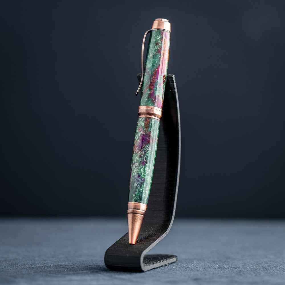 A handmade green, purple, and copper resin ballpoint pen on a black stand against a black background.