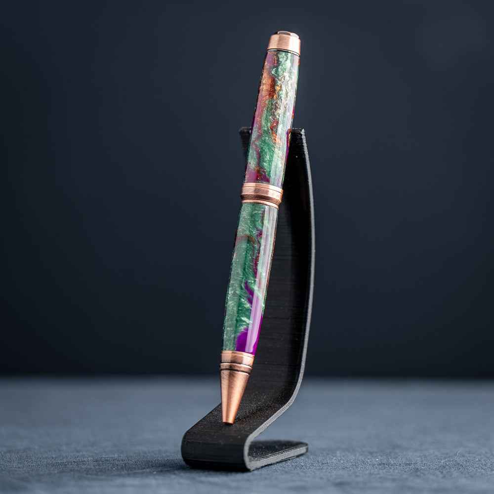 A handmade green, purple, and copper resin ballpoint pen on a wooden stand against a black background.