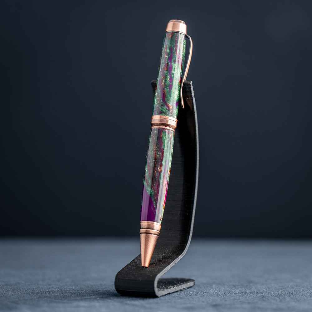 A handmade green, purple, and copper resin ballpoint pen on a wooden stand against a black background.