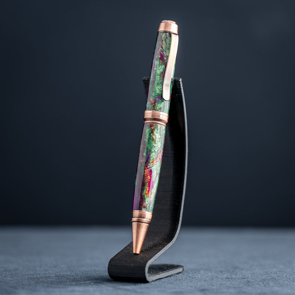 A handmade green, purple, and copper resin ballpoint pen on a wooden stand against a black background.