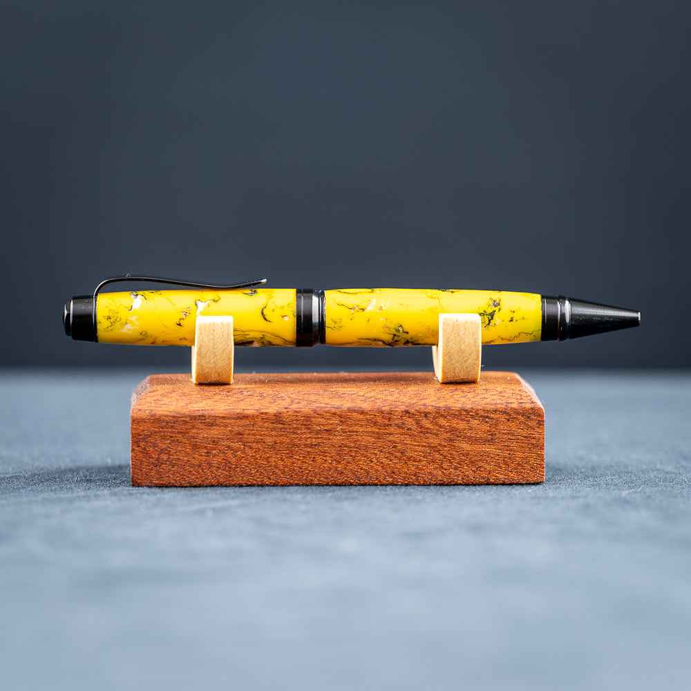 A handmade black, yellow, and white resin ballpoint pen on a wooden stand against a black background.