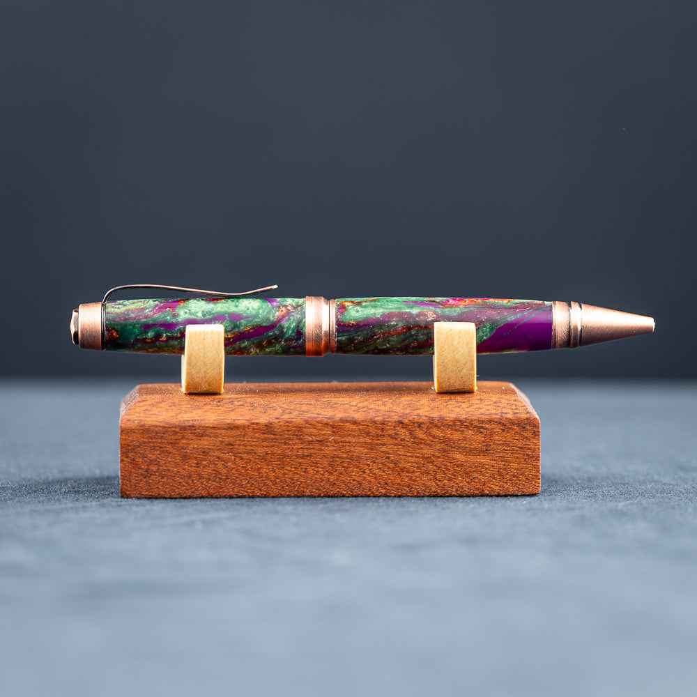 A handmade green, purple, and copper resin ballpoint pen on a wooden stand against a black background.
