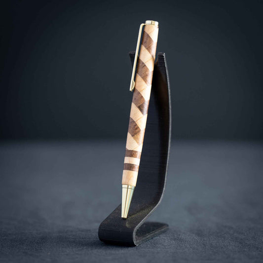 A handmade walnut and maple wood ballpoint twist pen with gold plating on a dark stand against a dark background.