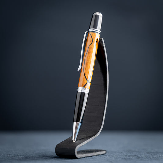 Handmade Osage orange wood and black wood ballpoint pen on a black stand against a dark background.
