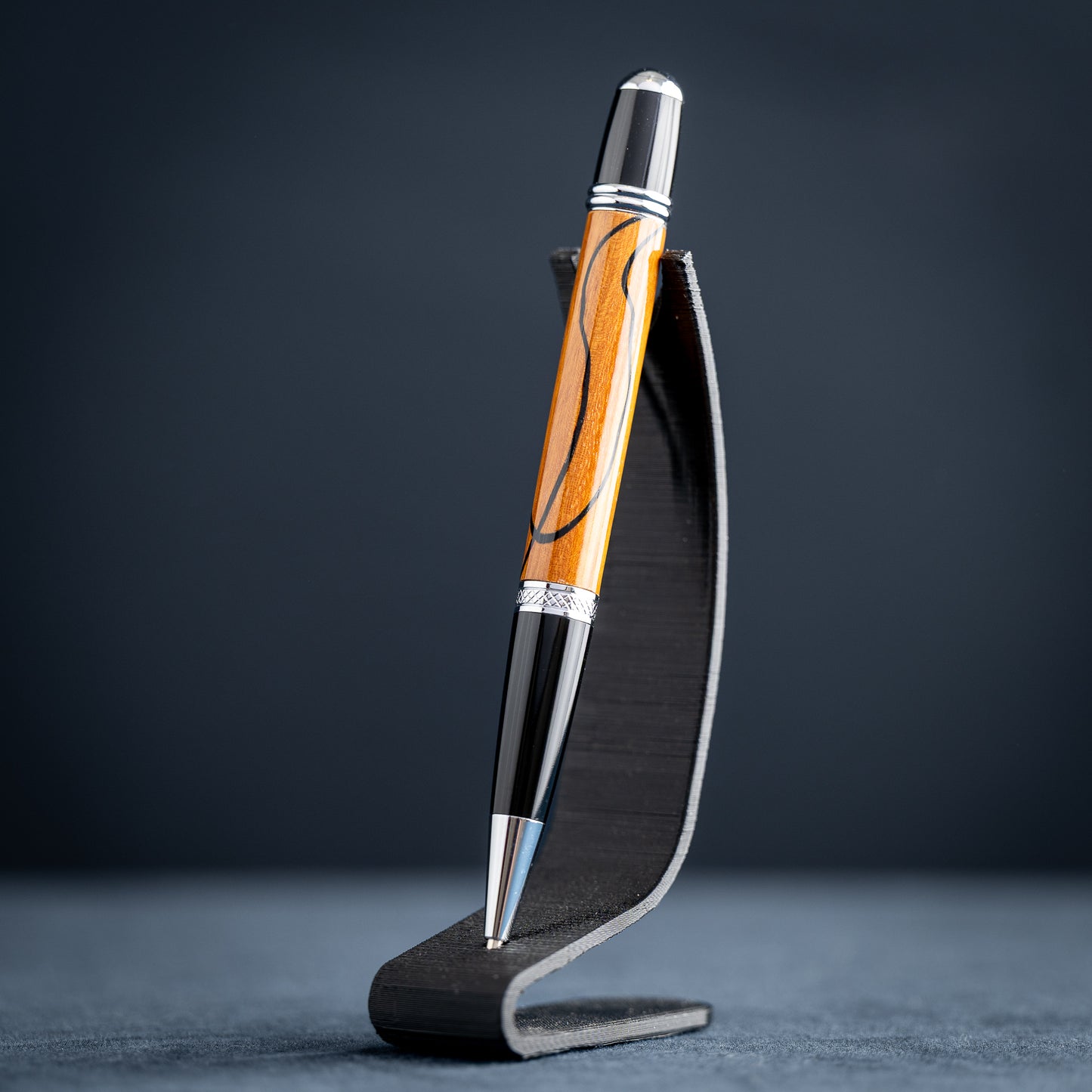 Handmade Osage orange wood and black wood ballpoint pen on a black stand against a dark background.