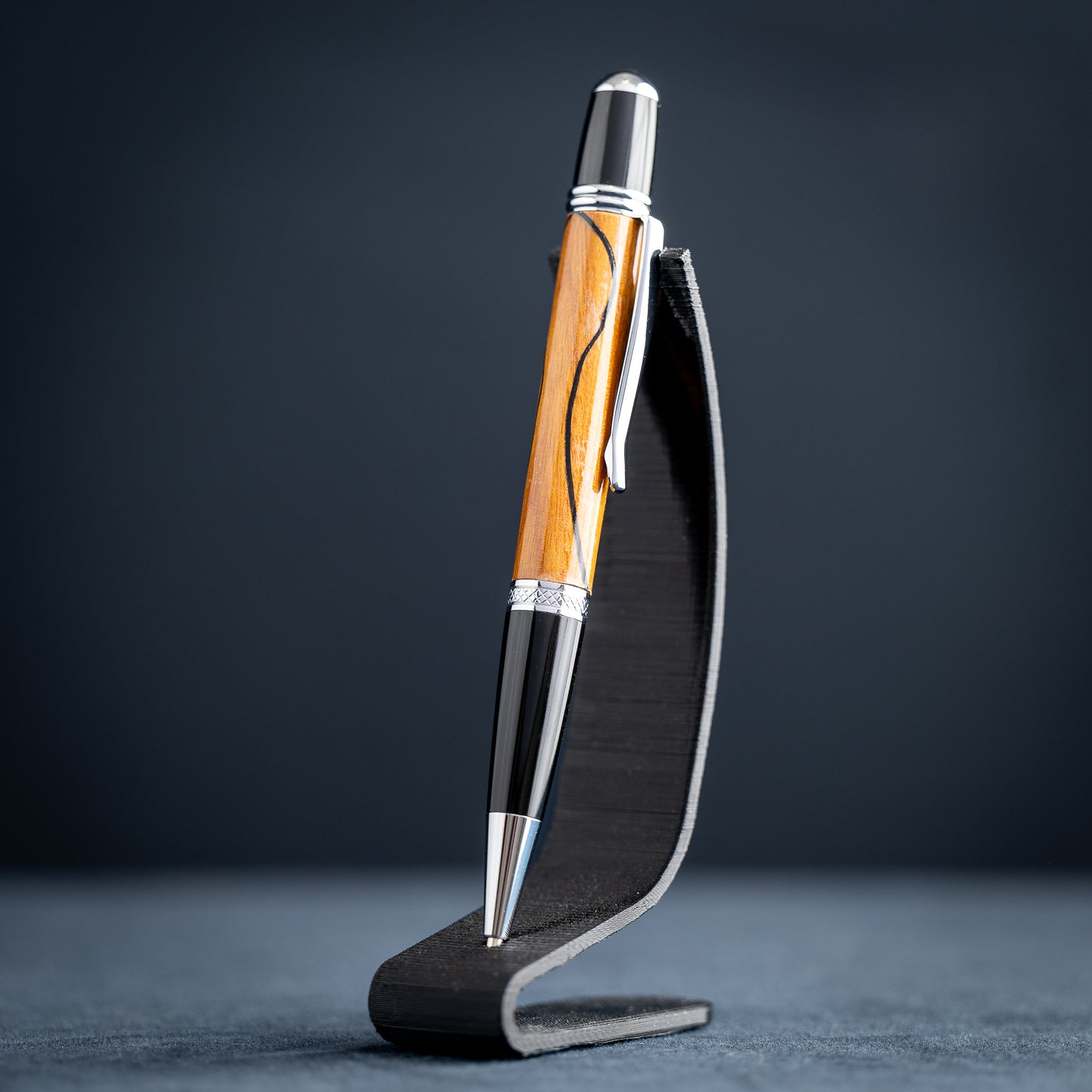 Handmade Osage orange wood and black wood ballpoint pen on a black stand against a dark background.